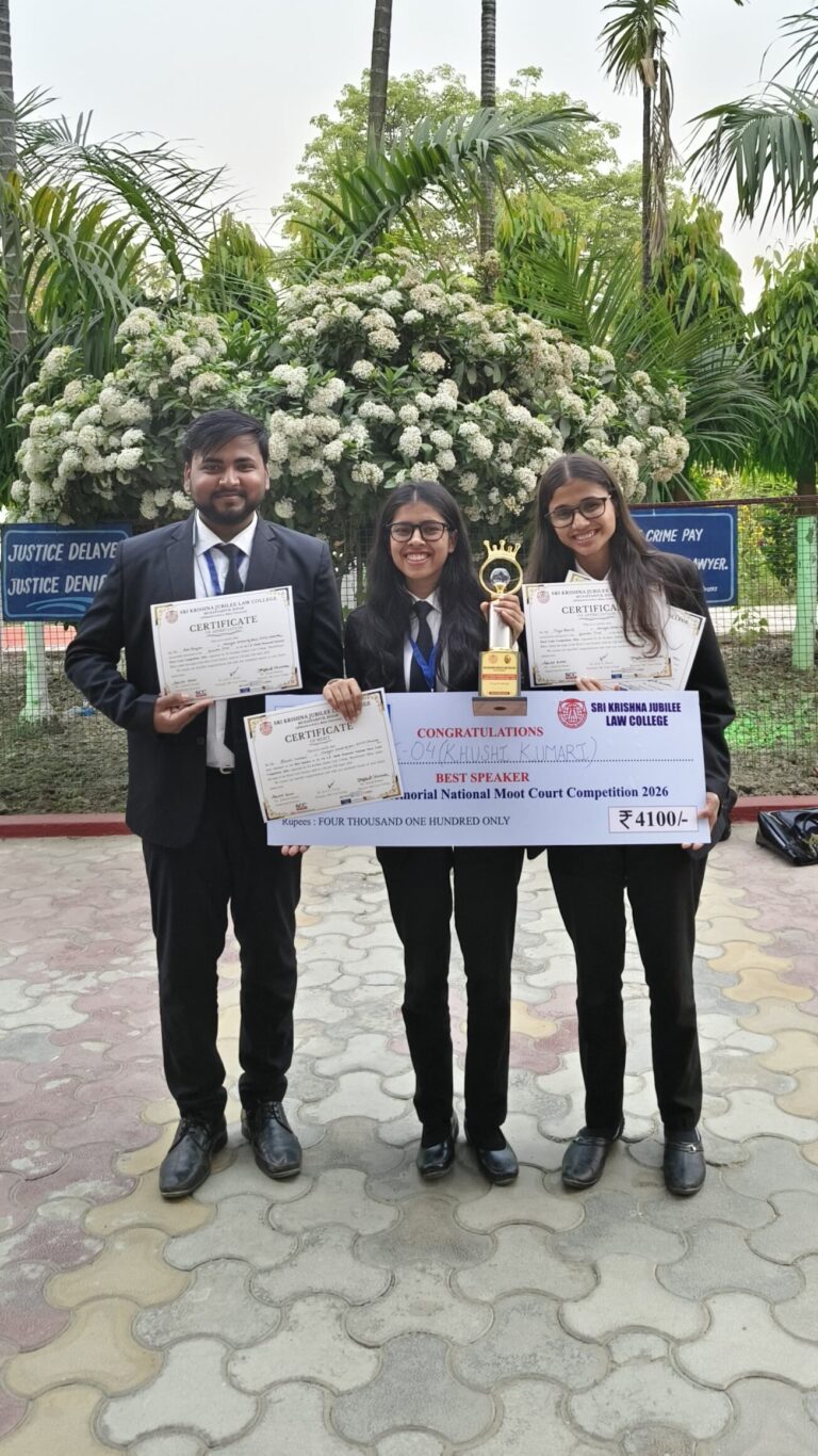 Three smiling students in suits pose with certificates, a trophy, and a large prize check outdoors in front of white flowering bushes.
