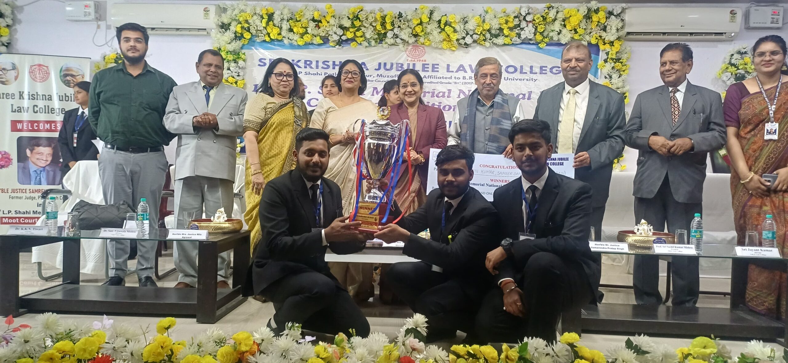 Group of formally dressed people posing with a large trophy on a flower-decorated stage at a law college award ceremony.