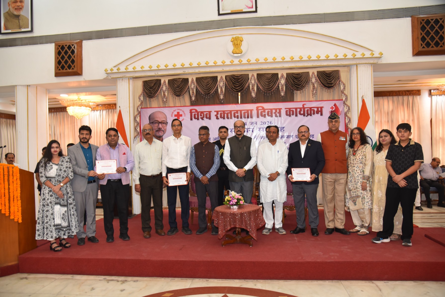Group of people on a stage posing for a photo at a formal event, several holding certificates, with a banner and Indian flags behind them.