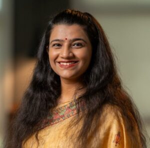 Portrait of a smiling woman with long dark hair wearing a gold saree and red bindi, facing the camera.