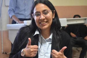Smiling woman with glasses giving two thumbs up in a blazer and striped shirt, indoors at a classroom or office setting.
