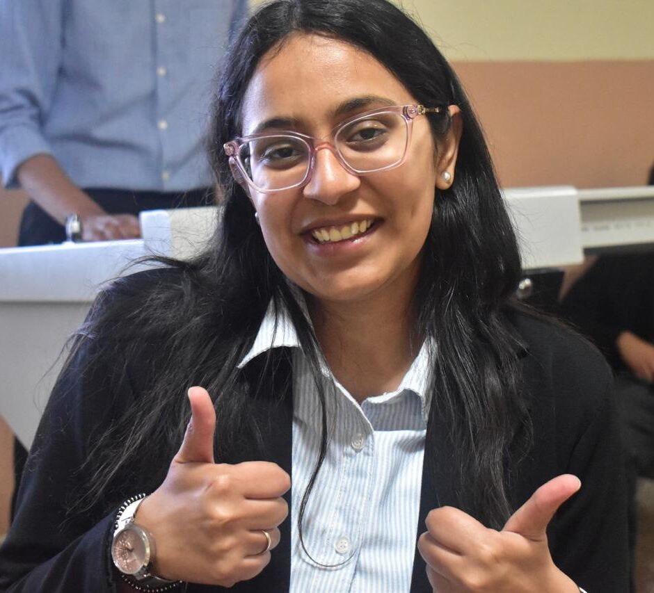 Smiling woman with glasses giving two thumbs up in a blazer and striped shirt, indoors at a classroom or office setting.
