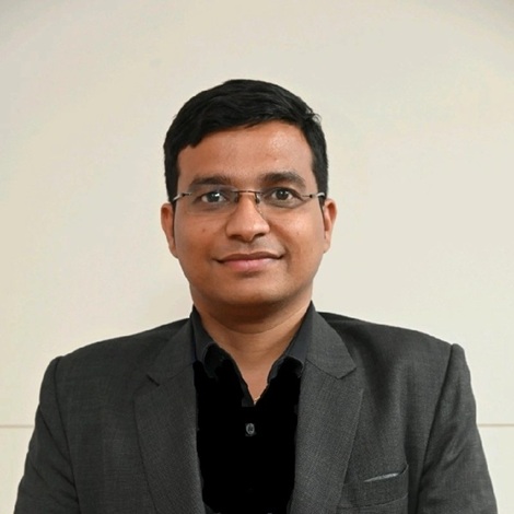 Portrait of a man in a dark blazer and black shirt, wearing glasses, facing the camera against a light background.