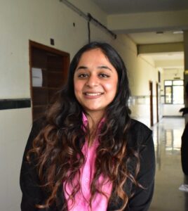 Smiling woman with long wavy hair wearing a pink shirt and black blazer in a hallway.