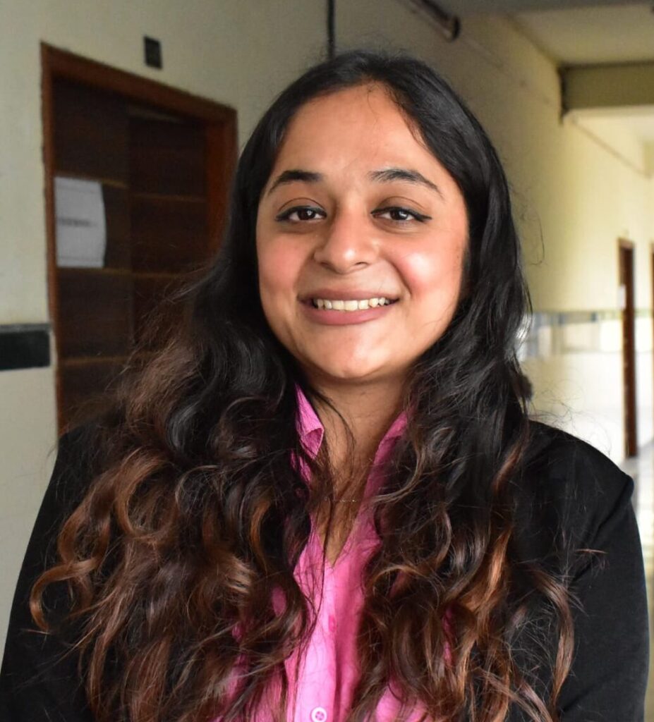 Smiling woman with long wavy hair wearing a pink shirt and black blazer in a hallway.