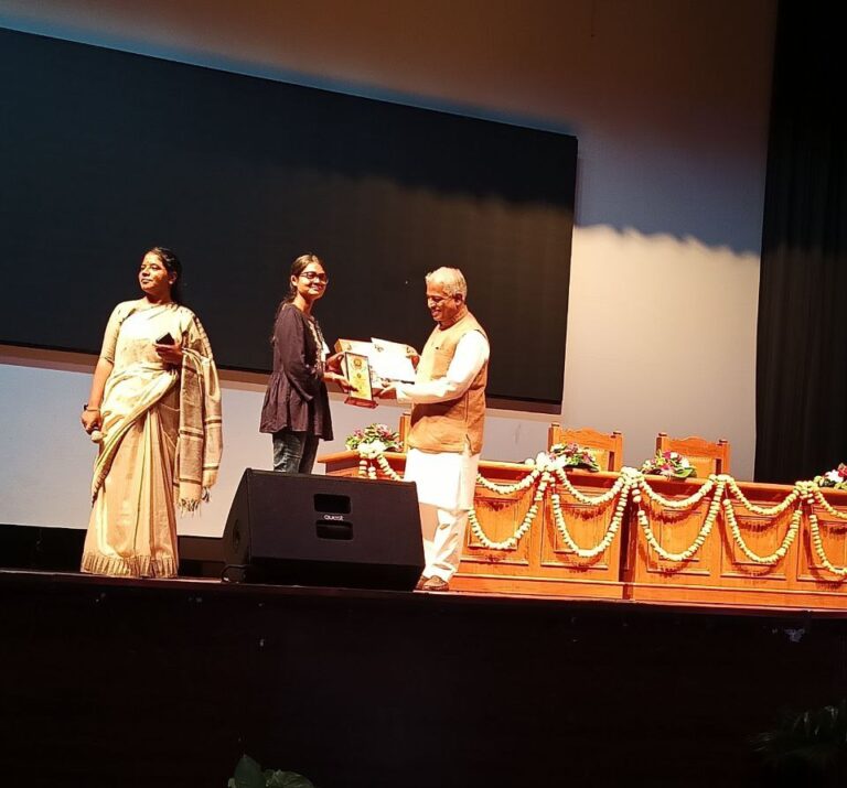 Three people on a stage during an award ceremony: a woman in a beige saree on the left, a recipient woman in the center receiving a certificate, and an older man in a vest handing the award. Garlands decorate the front of the stage.